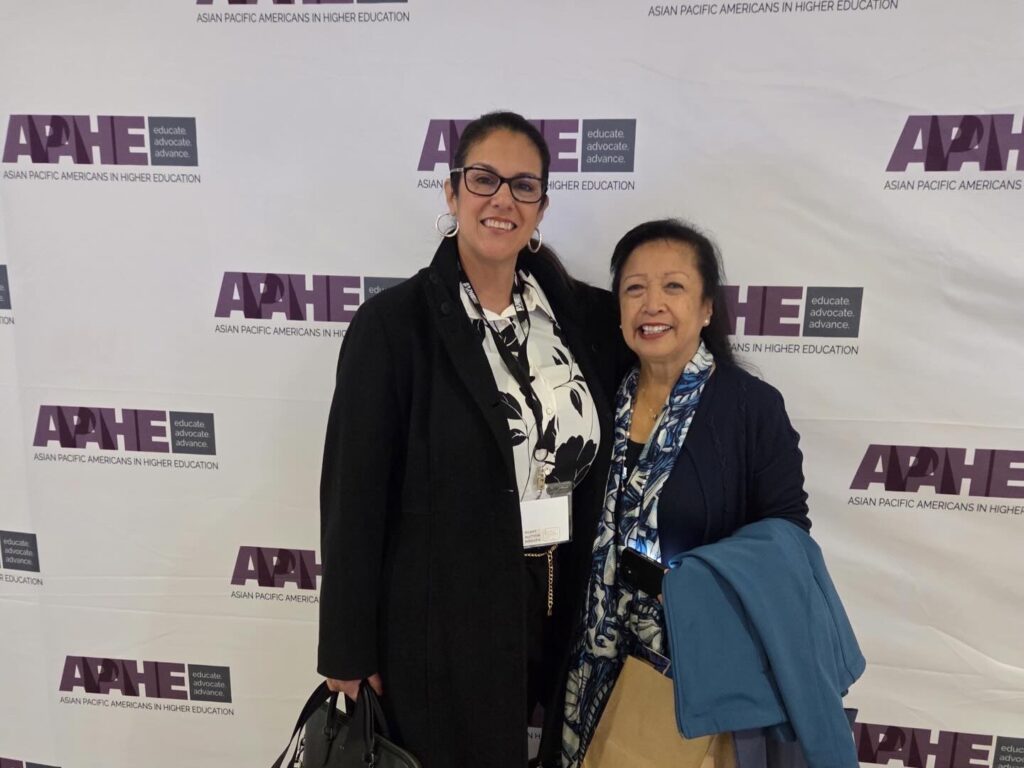 Two APAHE members standing in front of a APAHE Conference 2026 backdrop.