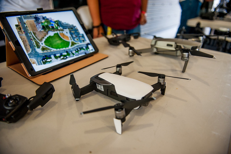 A compact multirotor drone sits on a display table next to a digital tablet displaying a high-resolution, stitched aerial map of a park, demonstrating drone mapping and data processing.