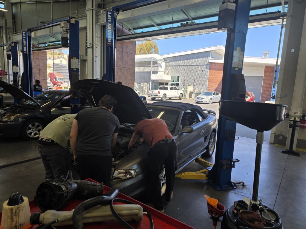 Three automotive technology students work together under the open hood of a car on a hydraulic lift inside the brightly lit Palomar College training shop.