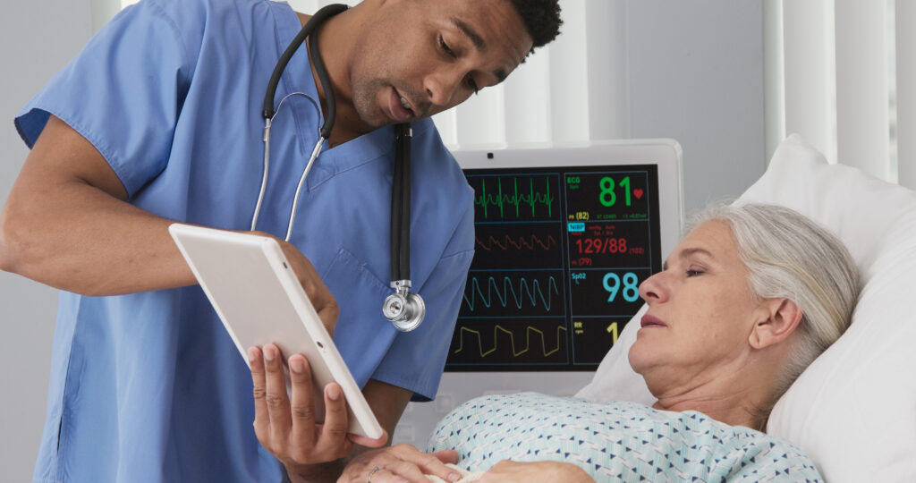 A male nurse in scrubs reviewing medical data on a tablet with a hospitalized patient next to a vital signs monitor.