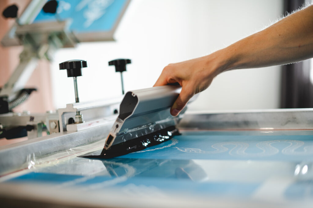 Close-up of textile screen printing equipment showing a hand manually pressing a squeegee to transfer ink onto a garment.