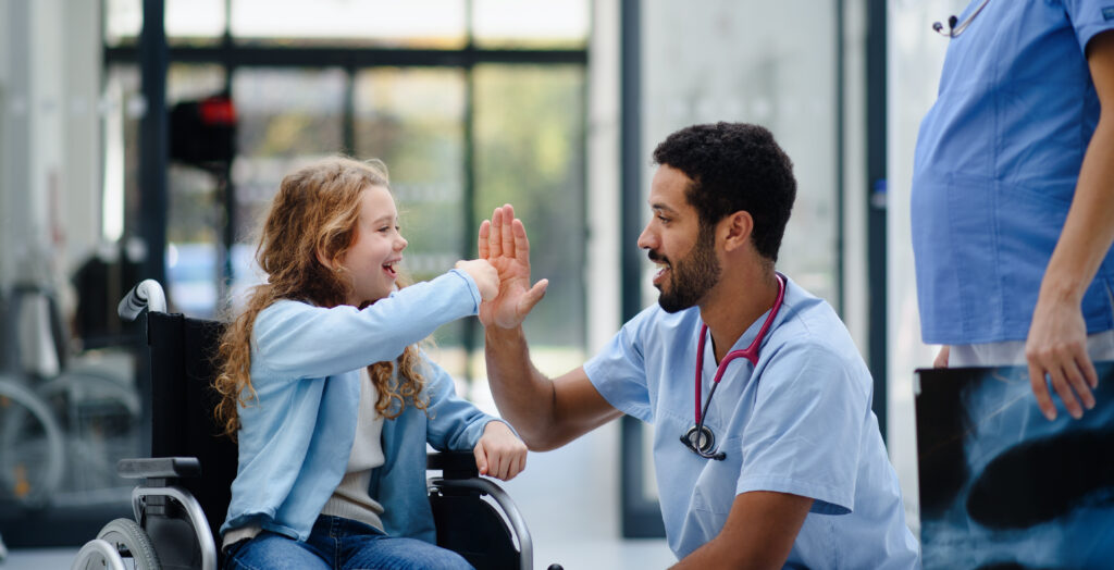 A male medical professional high-fiving a smiling young girl in a wheelchair.