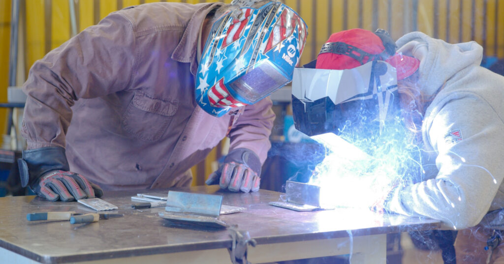 A welding instructor closely guiding a student as they work with a bright, sparking welding torch at a metal workbench.