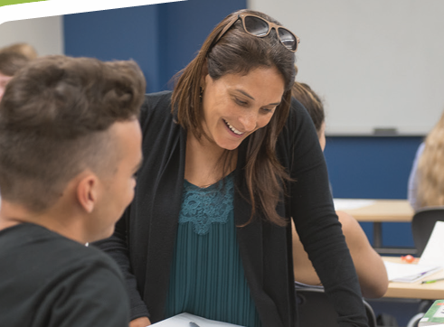 A smiling female instructor or career mentor leans forward to interact with a male student sitting at his desk in a classroom setting, showcasing active engagement and personalized learning.