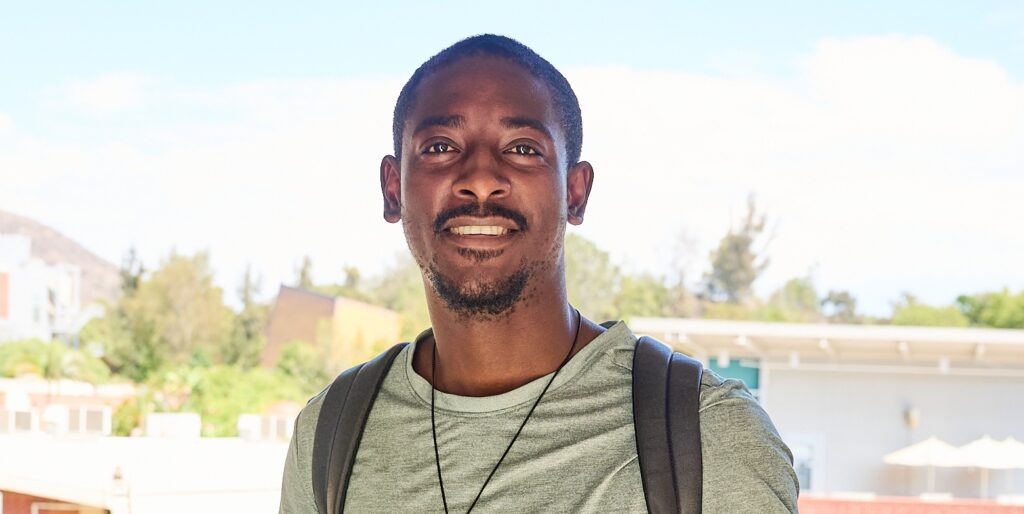 Smiling man stands outside on a balcony with college campus in the background.