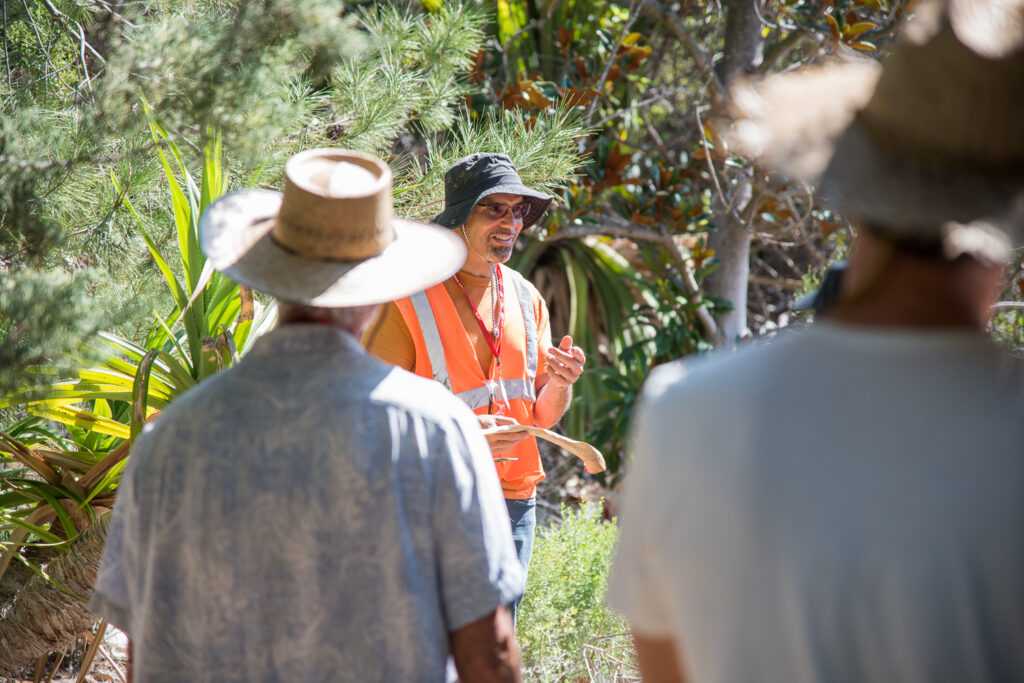 A man wearing an orange vest and a black bucket hat speaks to a group in front of a green background filled with plants.
