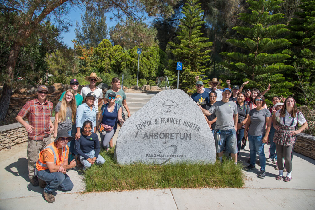 A group poses around a large stone monument that reads: Edwin and Frances Hunter Arboretum Palomar College. Large green trees are in the background against a blue sky.