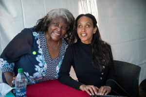 Haben Girma, a Black woman with long black hair, wearing a black long-sleeve dress smiles next to a Black woman with grey and black hair, wearing a blue shirt, also smiling.