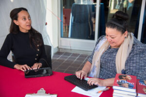 Haben Girma, a Black woman with long black hair, wearing a black long-sleeve dress, types on a keyboard alongside a woman with brown hair and olive skin and a blue blouse and tan scarf, who is smiling downwards while typing on a keyboard.