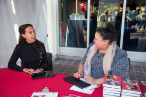 Haben Girma, a Black woman with long black hair, wearing a black long-sleeve dress, types on a keyboard alongside a woman with brown hair, olive skin and a blue blouse and tan scarf, who is smiling while typing on a keyboard.