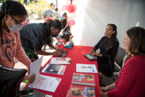 Haben Girma sits while a man wearing a black top types into her keyboard; they are at a table with a red tablecloth and papers and books on top. A woman wearing an orange shirt stands nearby, while a woman wearing a red dress sits smiling next to Haben Girma