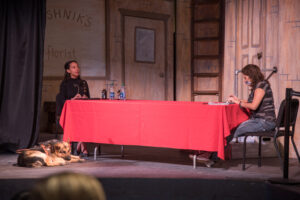 Haben Girma sits across a table with a red tablecloth while a student with brown hair and a dark top and jeans types on a keyboard across from her. Haben’s German Shepherd dog lays at her feet, the dog is brown and black and wearing a leather harness.