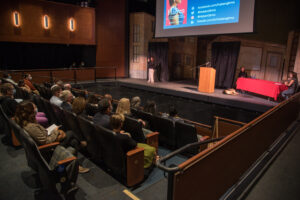 An audience looks on as Haben sits on stage across from a student at a long table with a red table cloth. A sign language interpreter stands signing in the corner of the stage.