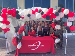 A group of people smile with Haben in the middle behind a table that has a red table cloth with ‘Palomar College’ written in white. They are surrounding by white, black, and red balloons under a white canopy.