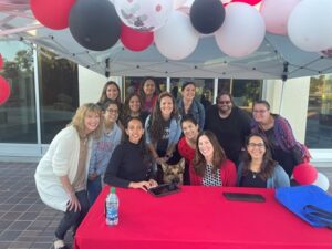 A group of people smile with Haben in the middle behind a table that has a red table cloth with ‘Palomar College’ written in white. They are surrounding by white, black, and red balloons under a white canopy.