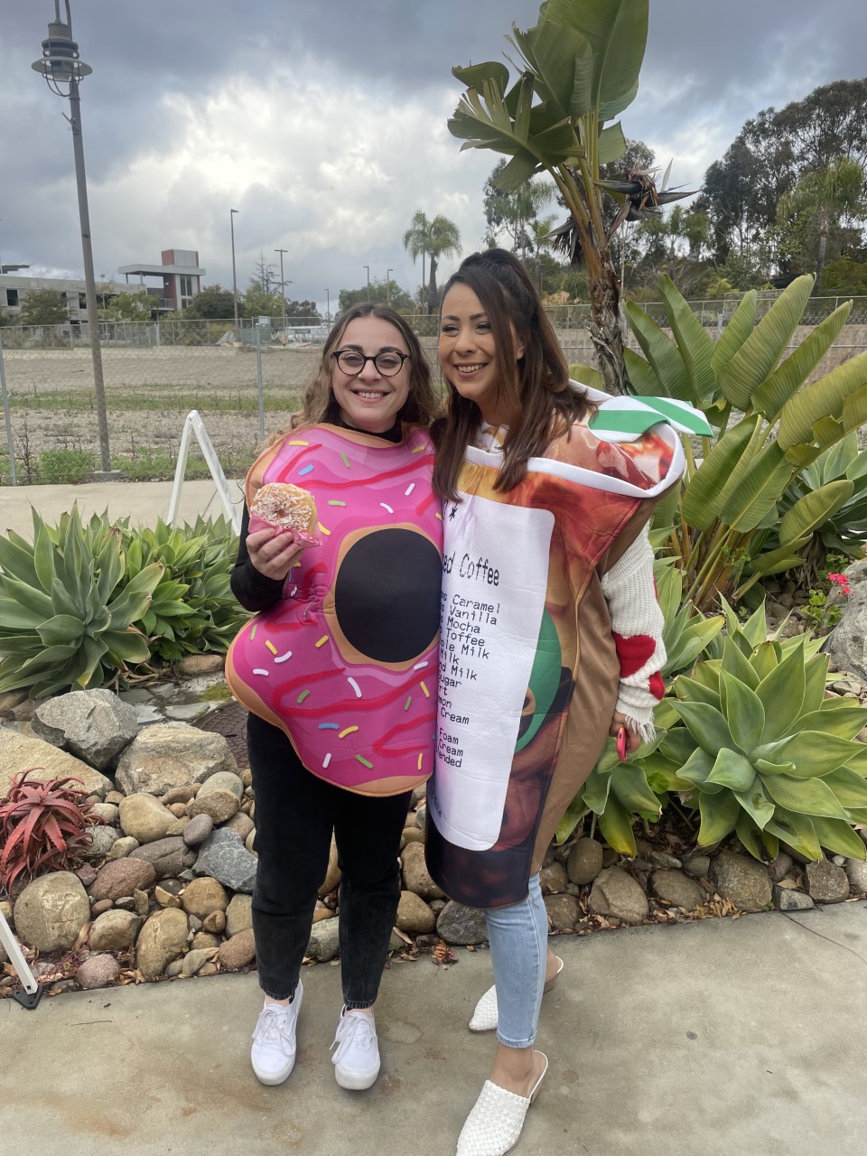 Two women wearing a donut costume and a coffee costume stand together smiling.