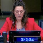 A woman with brown hair, wearing a bright red blazer, speaks at the United Nations. In front of her is a sign that reads CBM GLOBAL.