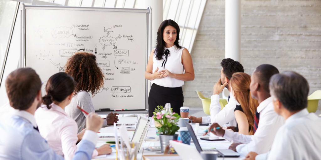 A lady talking in a meeting in the front of the room, explaining things on the whiteboard. 