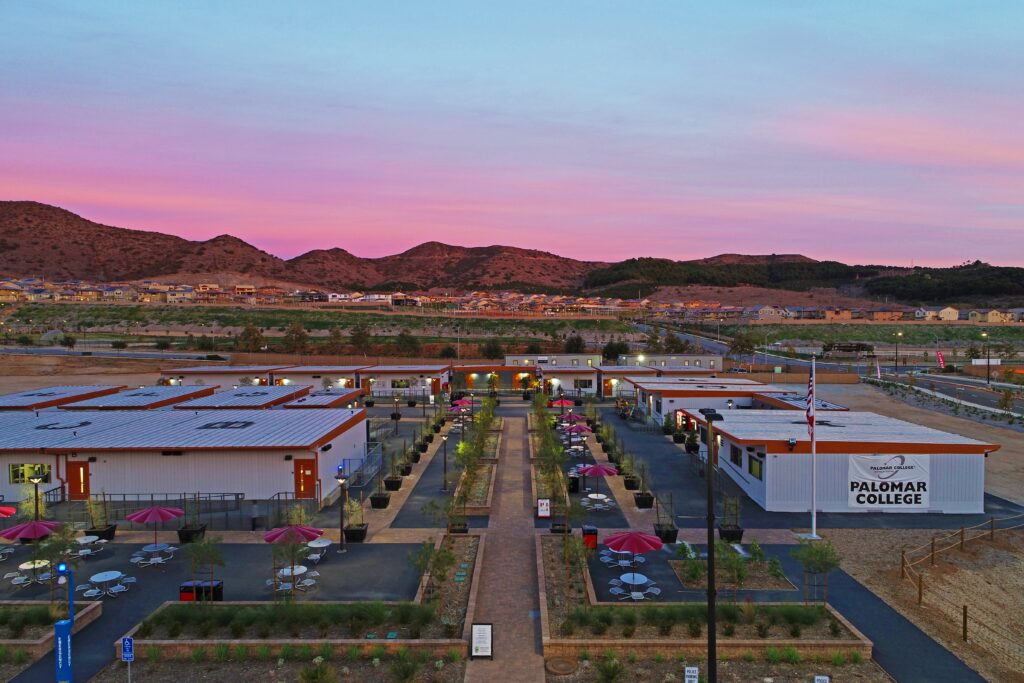 Panoramic view of Palomar College Fallbrook Campus at sunset with buildings, walkways, trees, and a backdrop of homes and hills under a pink-purple sky.