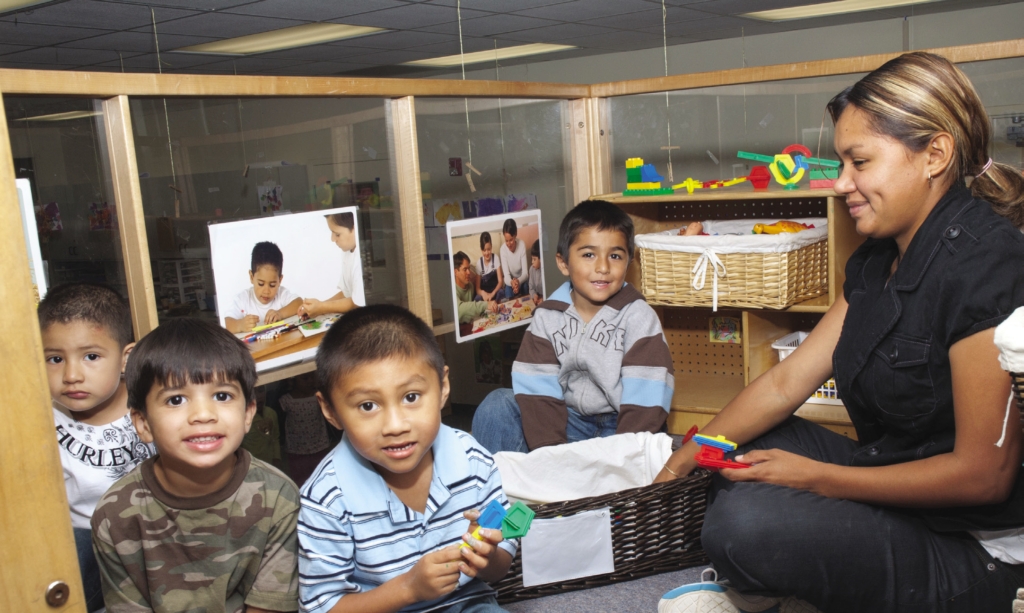 Several young children and an adult sit together in a classroom play area enclosed by low wooden partitions. The children are seated on the floor and hold colorful plastic building pieces, while the adult sits beside baskets of toys and learning materials. Photographs showing children engaged in activities are displayed on the partition walls behind them.