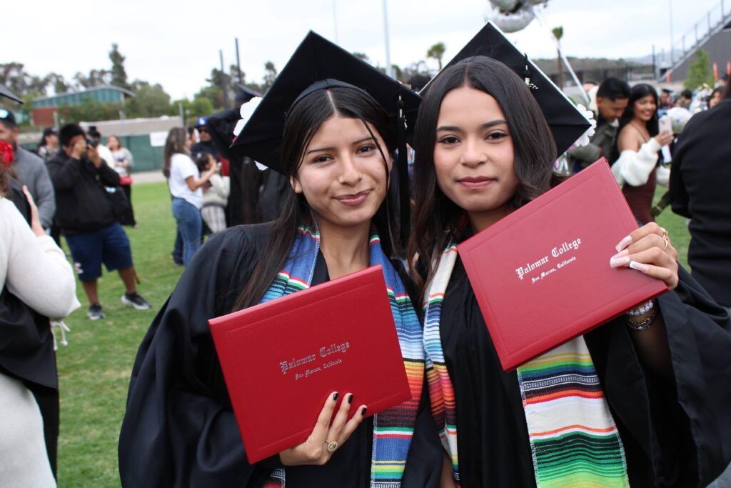 Two Bonsall High students in caps and gowns hold red Palomar College diploma covers during an outdoor graduation ceremony.