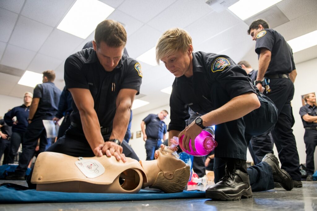 Palomar EME students practice CPR and ventilation on a training mannequin, with one person performing chest compressions while another uses a bag-valve mask in a classroom setting.