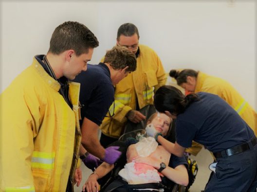 Several emergency response trainees and instructors in yellow jackets and dark uniforms practice a medical rescue scenario indoors. A participant is seated in a wheelchair while multiple responders provide care, including administering oxygen and assessing injuries.