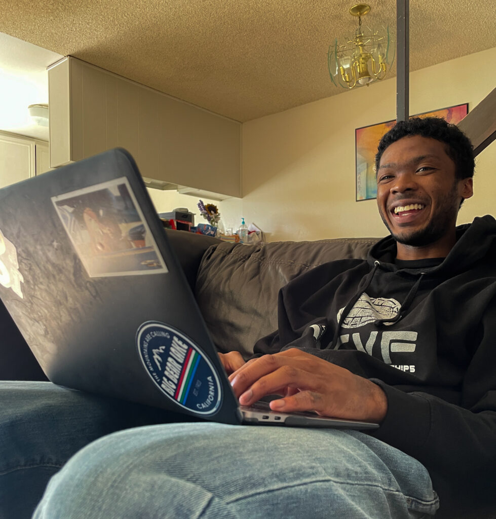 UCSD student Ben smiles at the camera while he works on his laptop on a black leather couch.