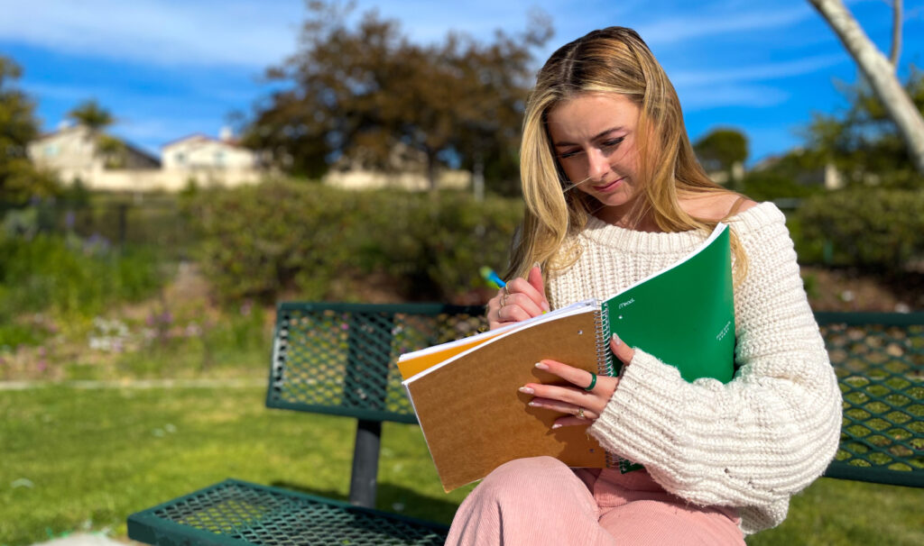 Female student sits on a green metal bench writing in her notebook on a sunny day.