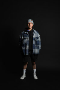 Male fashion model in gray and blue checkered flannel, black shirt, and black shorts.