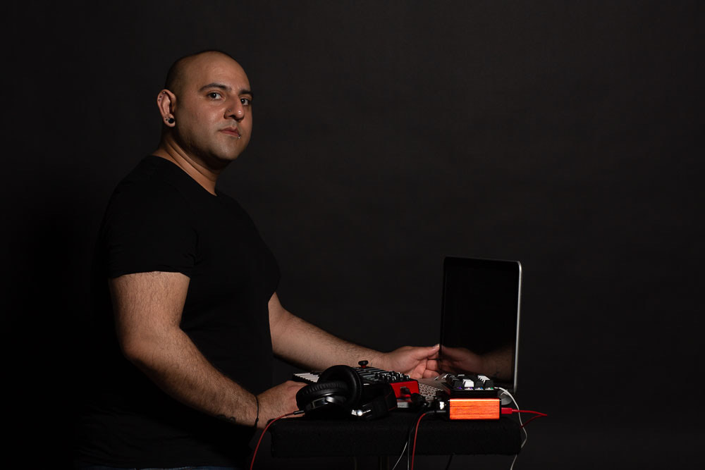 Zeshaun Hassan stands with his laptop and music setup with a black background.
