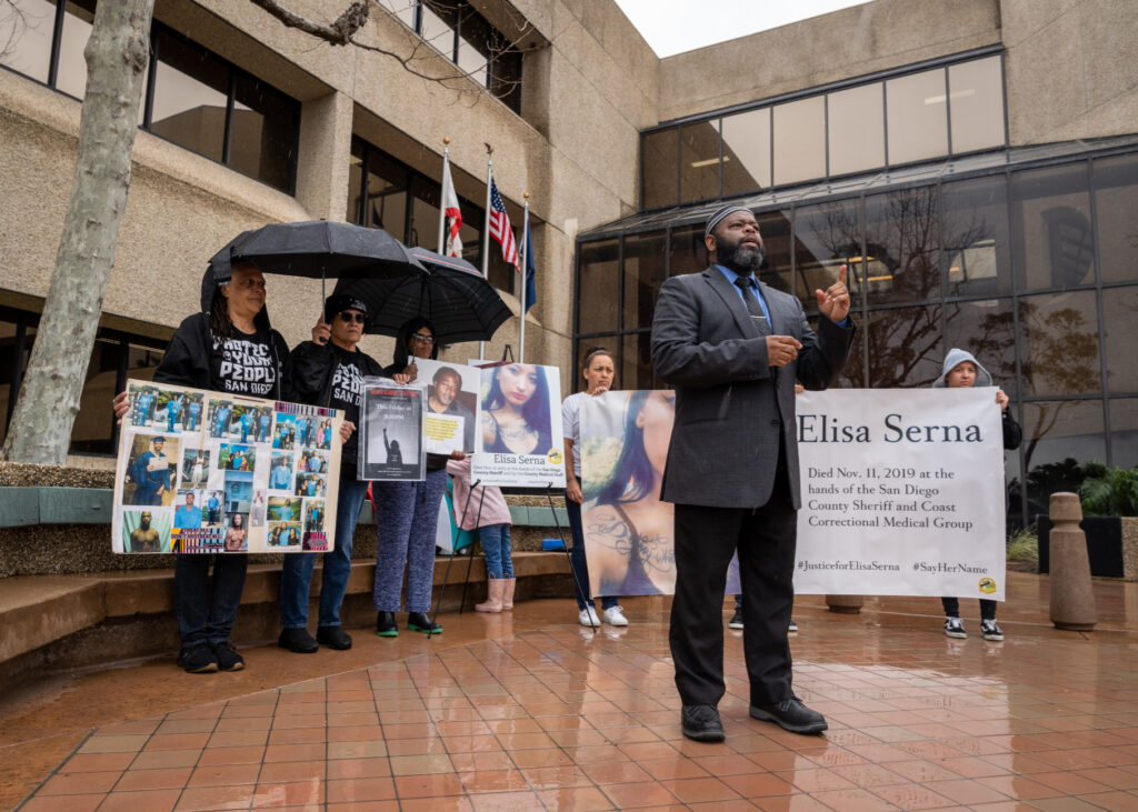 A group of people protests outside of the Escondido Police Department, holding posters and collages of those who died under custody. Yusef Miller from the North County Equity & Justice coalitions speaks during the protest.