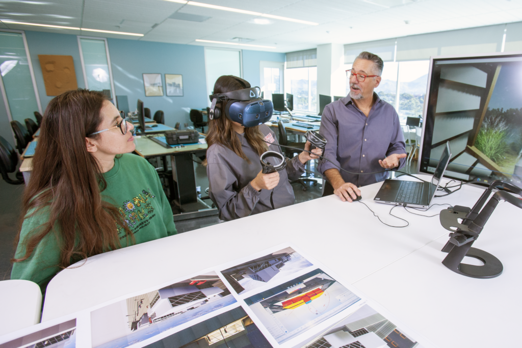 Two Architecture students, one watching the other test a virtual reality program. A faculty member instructs.