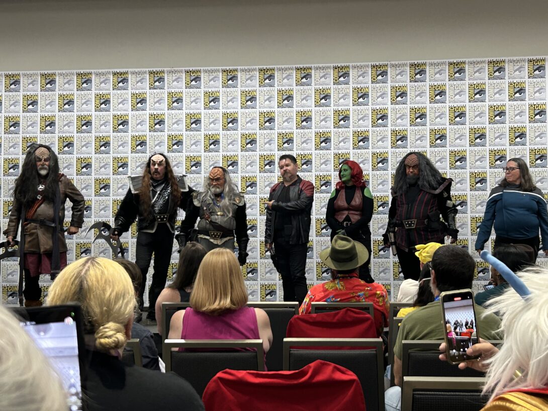 A group of people on stage. Four are dressed as Klingons, one is dressed as a captain from Star Trek, and another as a green alien. A woman is at the end in a blue shirt.