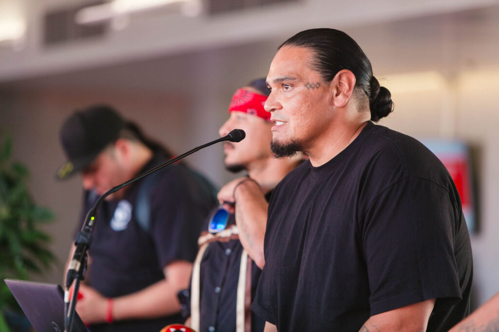 One Bird singer, Blue Eagle Vigil, greets attendees and speaks into a microphone indoors.