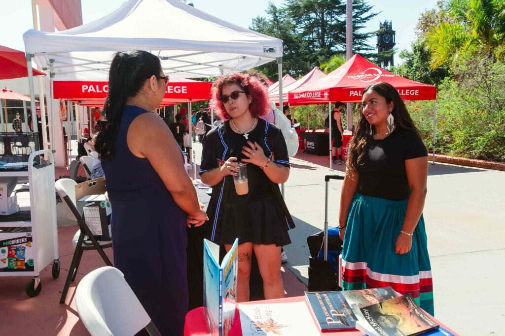 Individuals standing near tables with books and informational materials under red and white canopies.