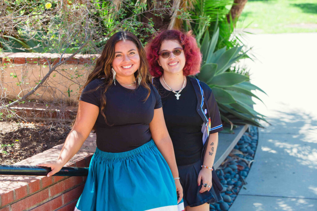 Two individuals standing outdoors, one wearing a blue ribbon skirt and the other in black attire.