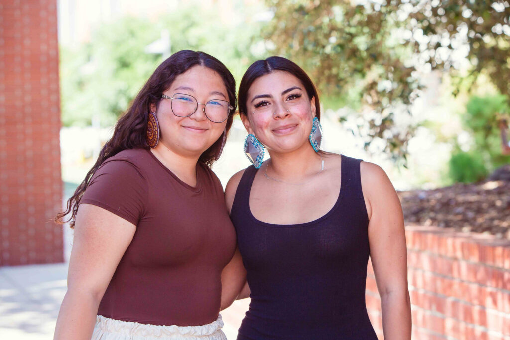 Two individuals standing near a brick wall wearing event attire and beaded earrings.