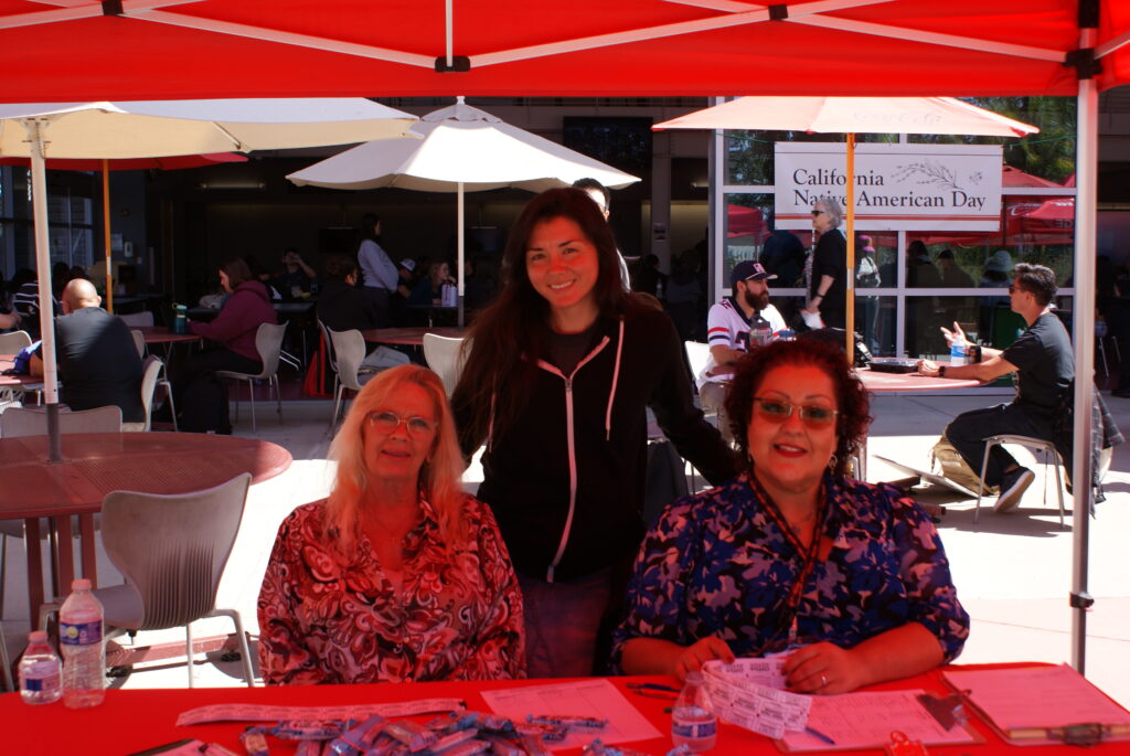 A table with two people seated and informational materials under a red canopy at the event.