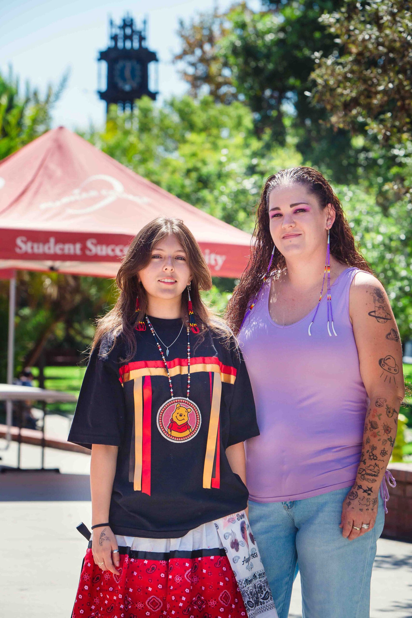 Cultural Celebration Outdoors Two individuals standing near a red canopy, one wearing a ribbon skirt and shirt, and the other in casual attire.