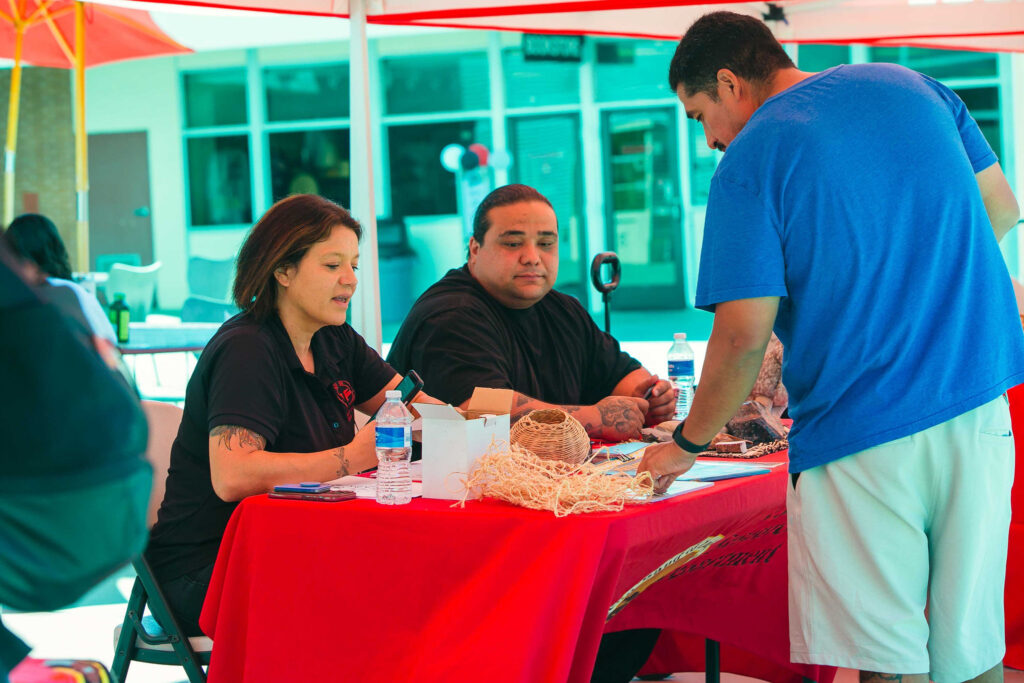 Two individuals seated behind a table with woven items and informational materials under a red canopy.
