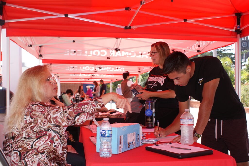 Attendees standing under red canopies at tables with bottled water, clipboards, and informational materials.