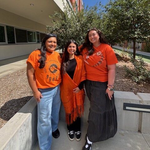 Three people in orange clothing stand outside a campus building near trees, supporting Every Child Matters Day.