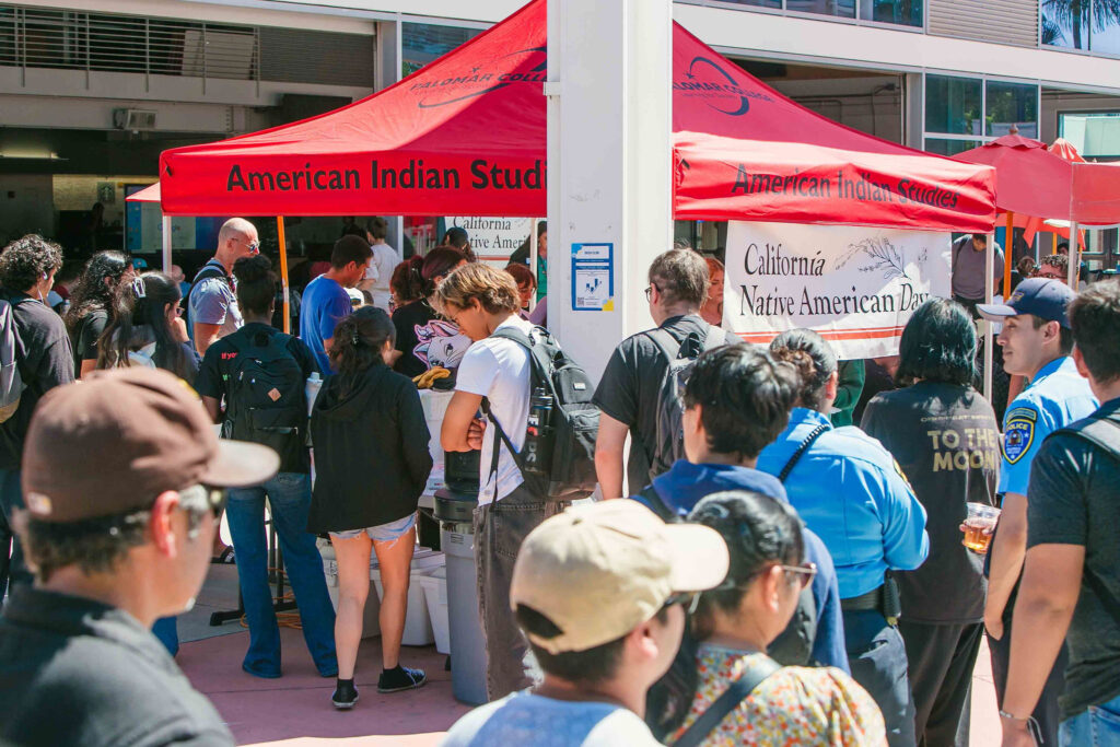 Large group of attendees gathered under red canopies near a banner reading 'California Native American Day.'