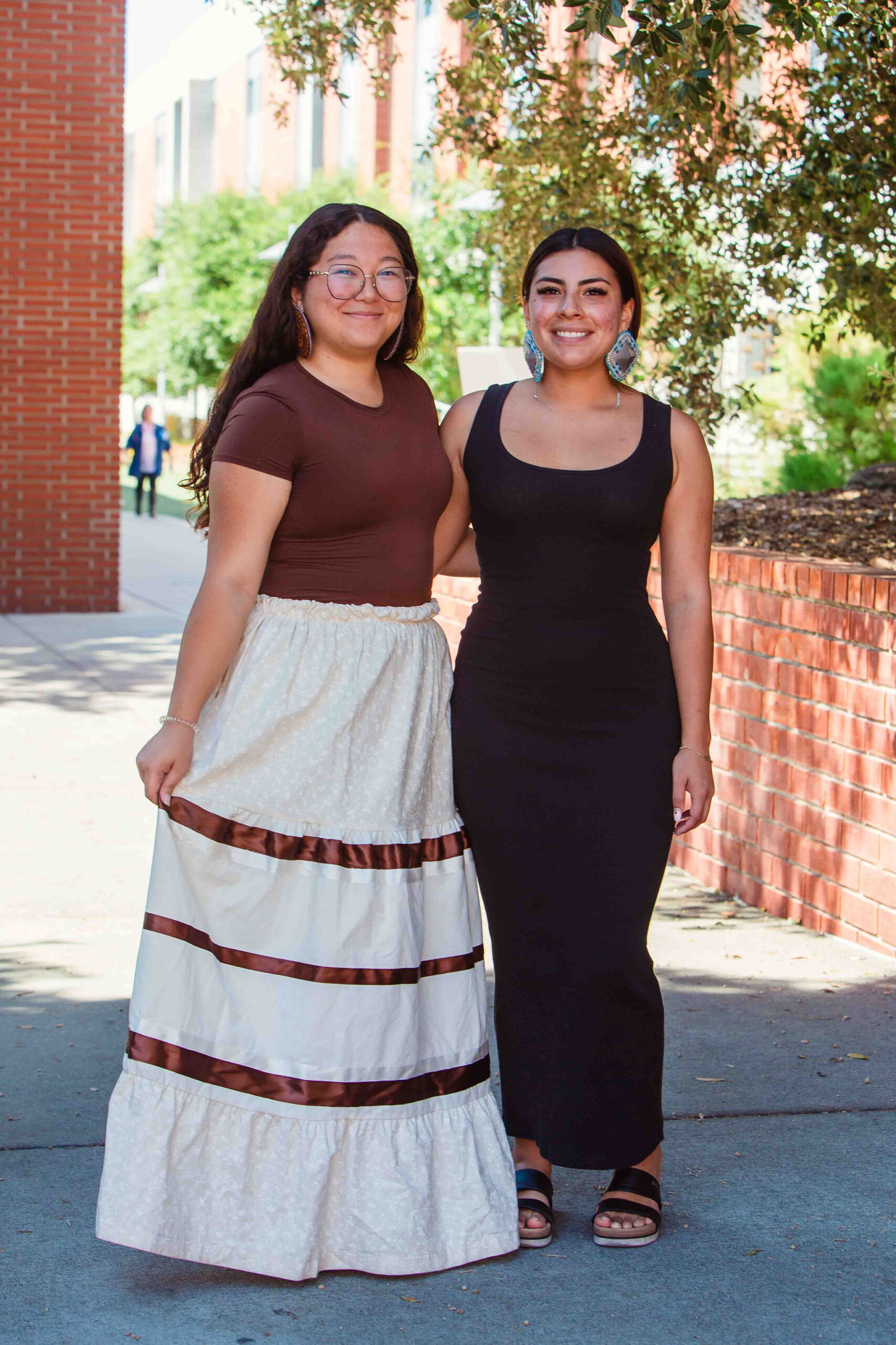 NSPIRE Students in Traditional Attire Two individuals standing outdoors, one wearing a cream ribbon skirt and the other in a black dress.