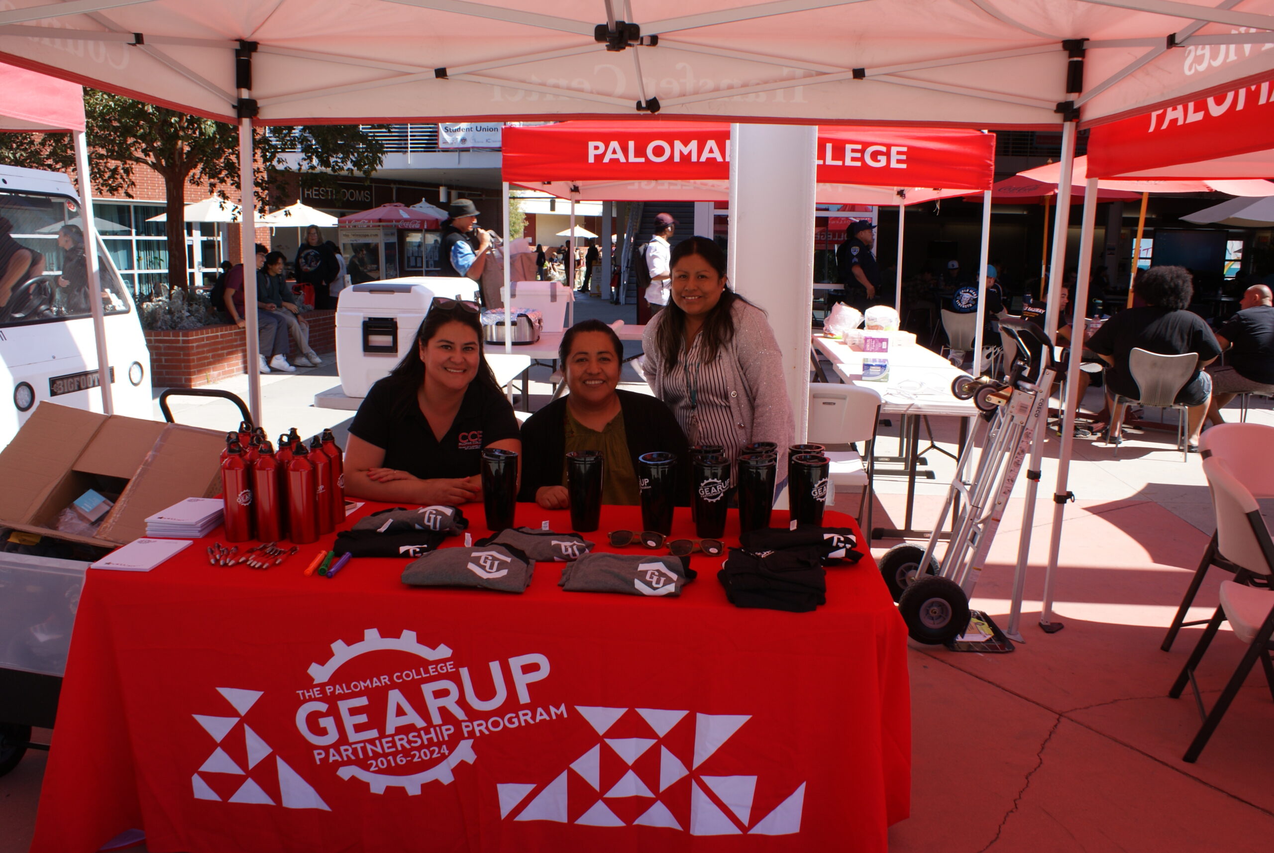 GEAR UP Partnership Program Table Table with a red GEAR UP Partnership Program cloth displaying water bottles, t-shirts, and informational materials under a canopy.