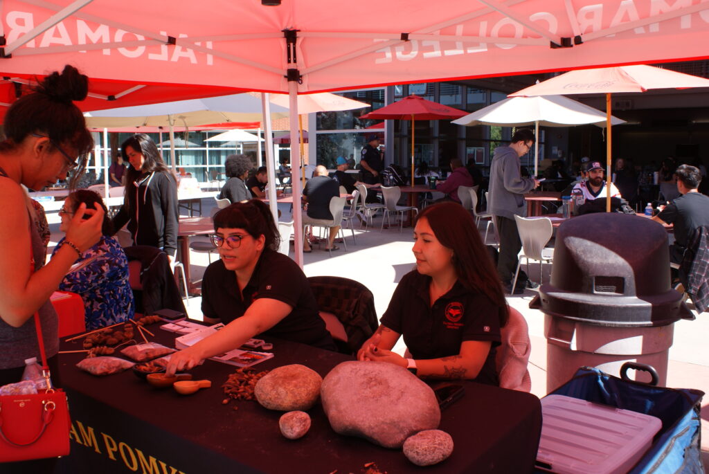 An attendee speaks with representatives at a table displaying traditional grinding stones and seeds under a red canopy.