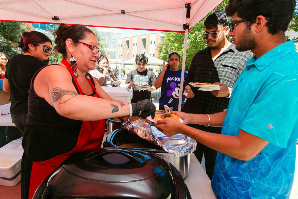 Individual serving food from trays under a red canopy with plates, napkins, and drinks visible on table.