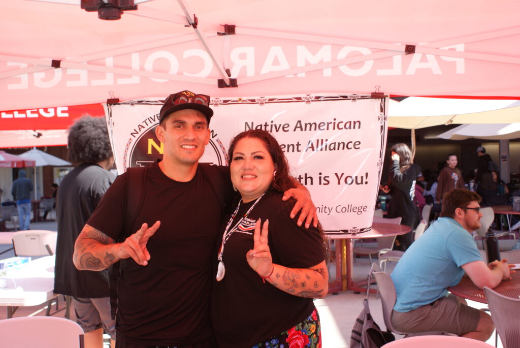 Two people standing under a red canopy in front of a Native American Student Alliance banner.