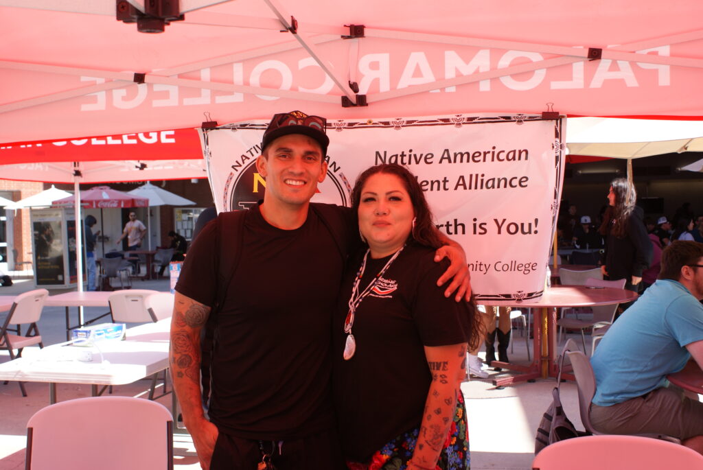 Two individuals posing under a red canopy in front of a banner promoting Native American Student Alliance.